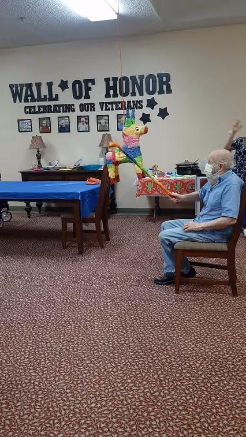 An elderly man wearing a mask sits on a chair indoors, hitting a colorful piñata shaped like a donkey with a stick. Behind him is a wall decorated with the words 'WALL OF HONOR CELEBRATING OUR VETERANS' and framed photos. The room has tables covered with blue and red tablecloths and a patterned carpet.