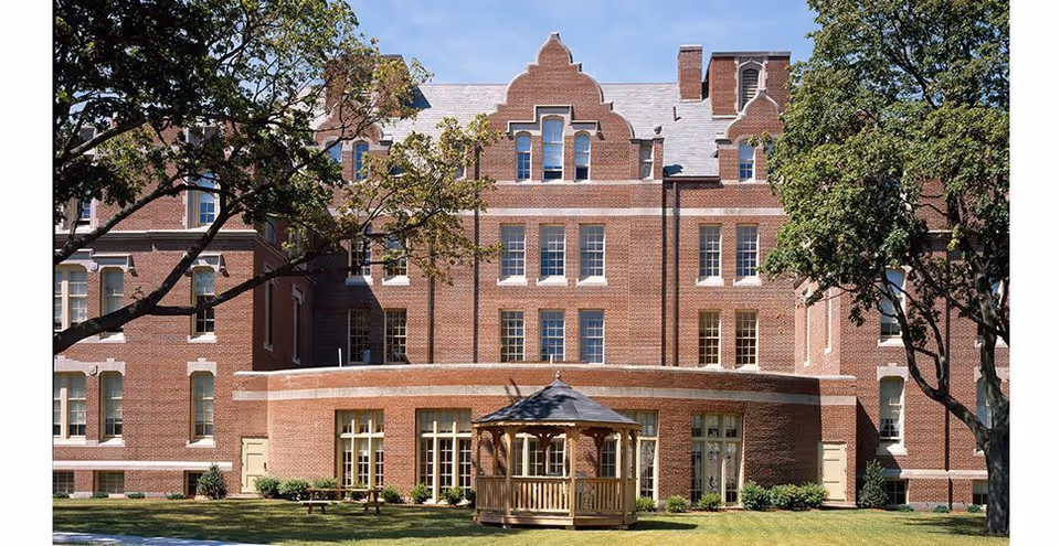 A large, multi-story brick building with numerous windows and decorative architectural details. In front of the building is a grassy area with a wooden gazebo and several trees providing shade.