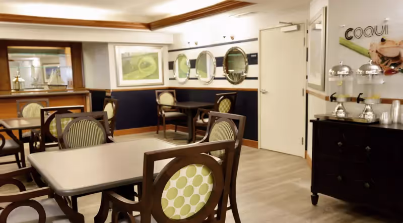 A dining area with several wooden tables and chairs featuring green and white patterned cushions. The room has light-colored walls with dark blue wainscoting, decorative mirrors on the wall, framed artwork, and a sideboard with beverage dispensers and cups. The floor is light wood, and the ceiling has recessed lighting with a wooden trim.
