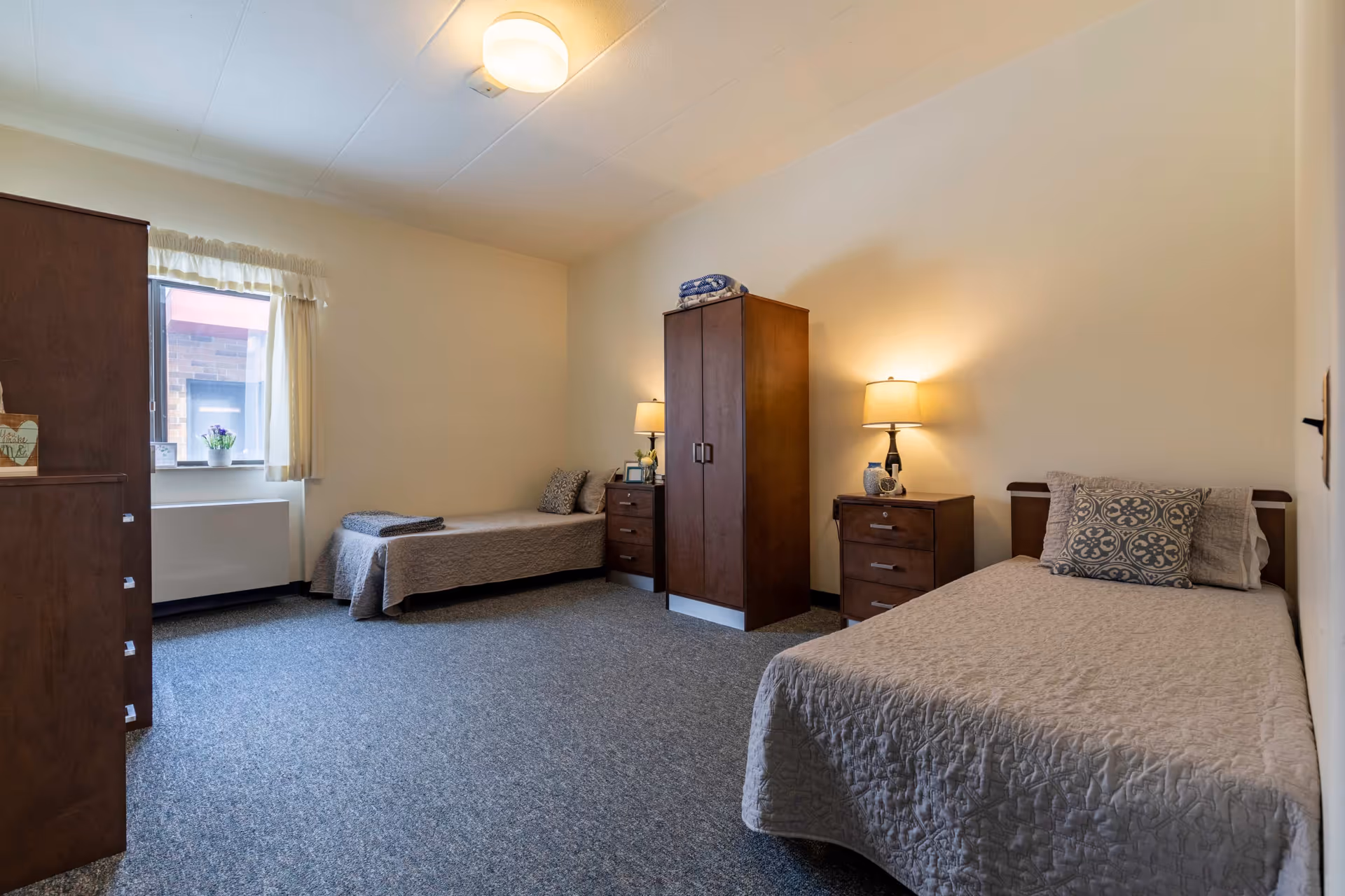A simple, clean bedroom with two single beds covered in beige quilts, each with a decorative pillow. Between the beds is a wooden wardrobe and two matching nightstands with lamps. A window with cream curtains lets in natural light, and the floor is carpeted in a blue-gray tone.