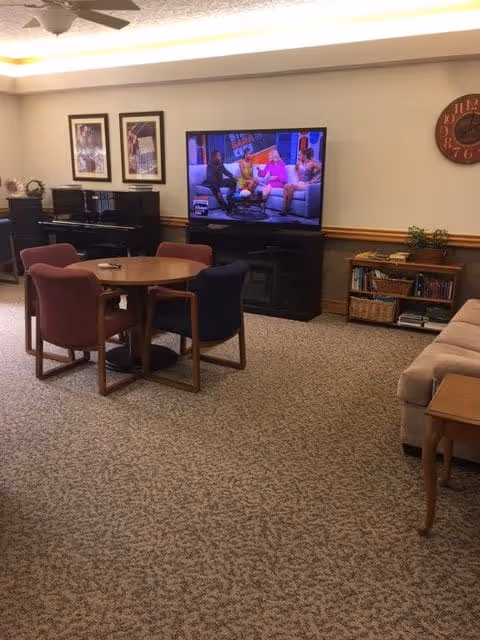 A cozy living room area with a round wooden table surrounded by four upholstered chairs in different colors. A large flat-screen TV is mounted on a black stand against the wall, displaying a talk show with four people sitting on a couch. The room has beige carpet, a beige sofa on the right, a wooden side table, a bookshelf with books and baskets, and framed pictures on the wall. A ceiling fan is visible above.