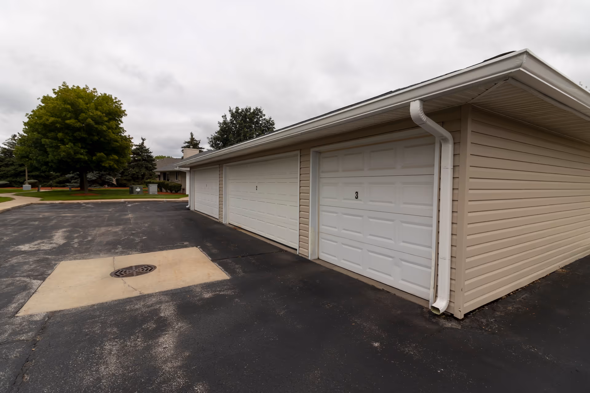 Exterior view of a beige building with three white garage doors numbered 1 and 3 visible, situated on a paved area with a storm drain. Trees and greenery are visible in the background under a cloudy sky.