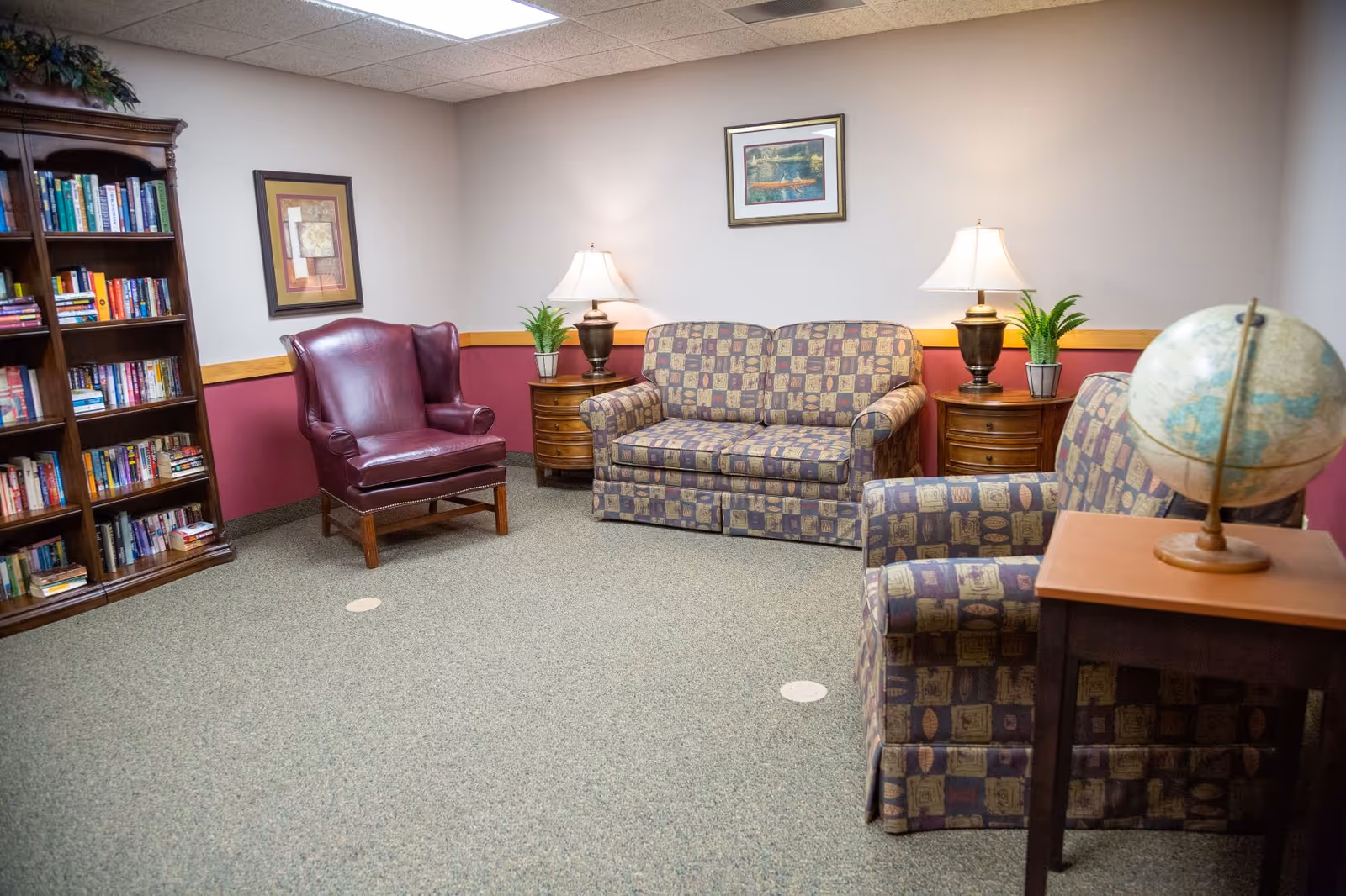 A cozy sitting area in a senior living facility with a patterned loveseat and armchair, a burgundy leather armchair, two wooden side tables with lamps and small potted plants, a wooden bookshelf filled with books, a framed picture on the wall, and a globe on a small wooden table.