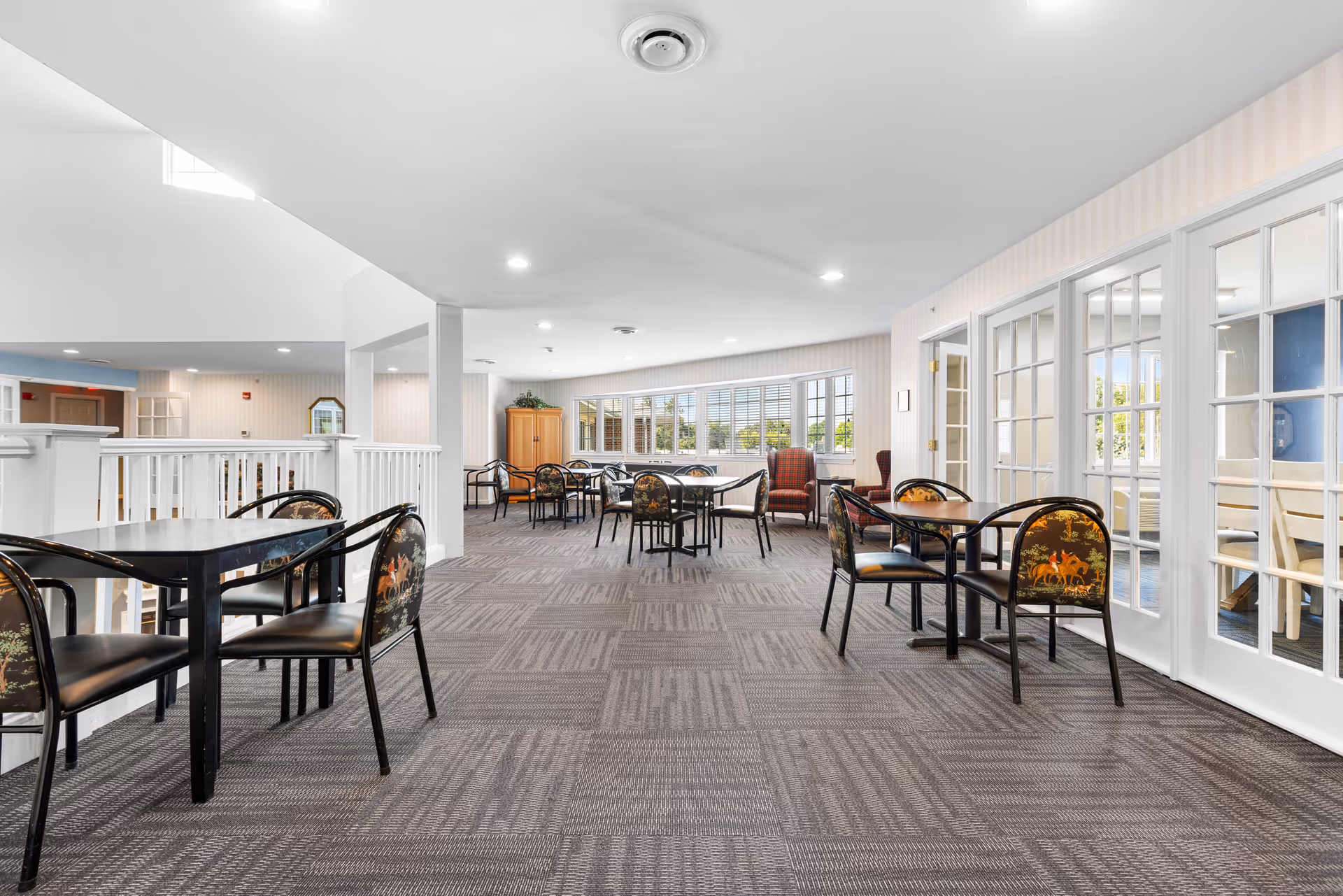 A bright and spacious common area in an assisted living facility with multiple tables and chairs arranged for seating. The room features large windows letting in natural light, patterned carpet flooring, and white walls with subtle vertical stripes. There are glass-paneled doors on the right side and a wooden cabinet with plants near the windows in the background.