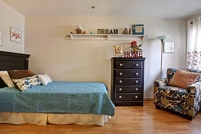 A cozy bedroom with a single bed covered in a teal quilt and several decorative pillows. Next to the bed is a dark wooden chest of drawers with decorative items on top, including a sign that says 'HOME' and a small plant. Above the chest is a white wall shelf with more decorative items and a sign that says 'DREAM'. To the right is a patterned armchair with a brown pillow, a tall floor lamp, and a window with white curtains featuring a leaf pattern. The room has light-colored walls and wooden flooring.