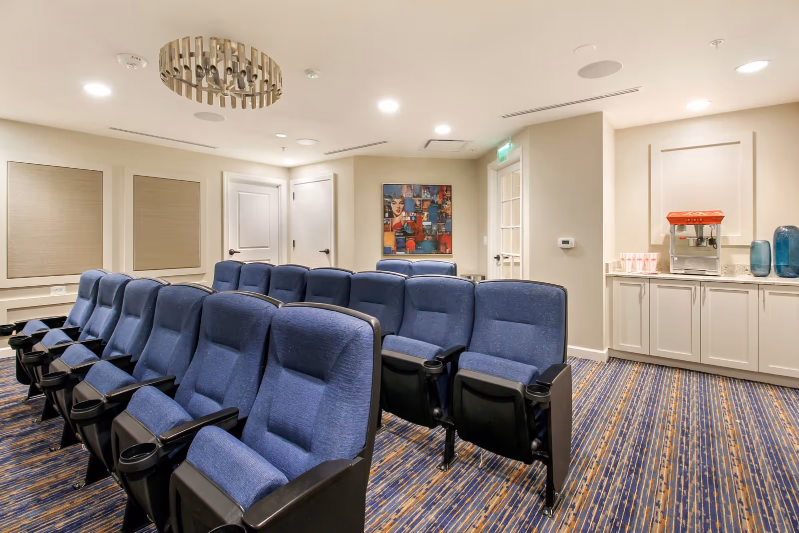 A small theater room with two rows of blue cushioned seats with black armrests and cup holders. The room has beige walls, a patterned carpet, a modern circular ceiling light fixture, and a countertop with a popcorn machine and cups on the right side. There is a colorful framed artwork on the far wall and two closed white doors.