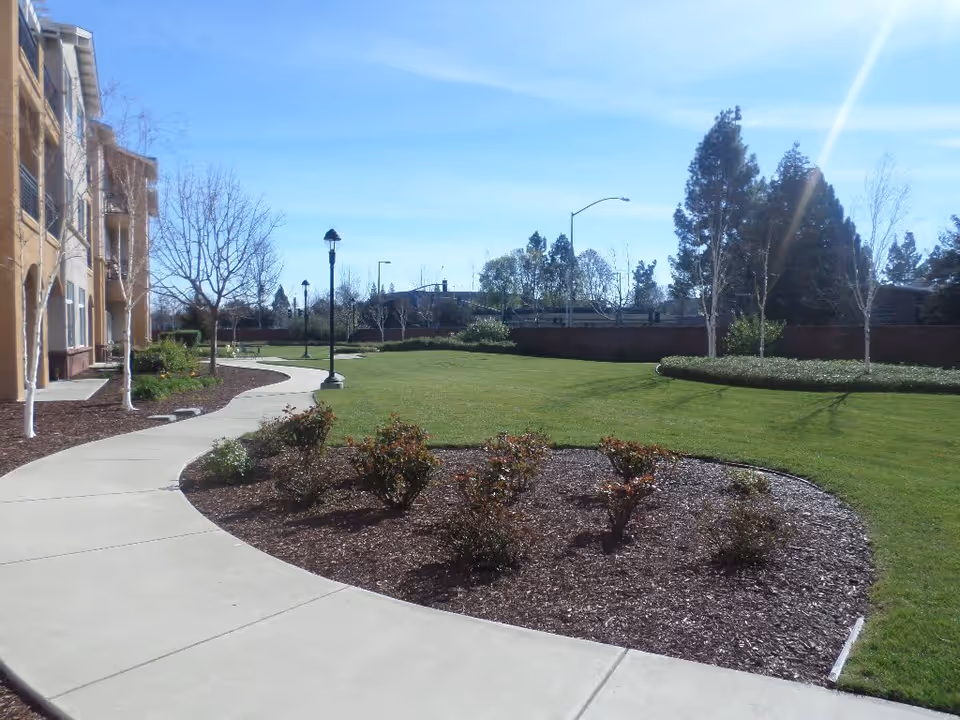 A sunny outdoor area at Heritage Estates featuring a curved concrete walkway bordered by landscaped flower beds with small bushes and trees. There is a grassy lawn area with more trees and lampposts, and a multi-story building is visible on the left side of the image.