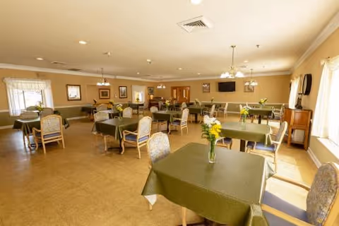 A spacious dining room with multiple tables covered in green tablecloths, each adorned with a vase of yellow flowers. The room has beige walls, several windows letting in natural light, framed pictures on the walls, and ceiling lights providing illumination. Chairs with patterned upholstery are arranged around the tables.