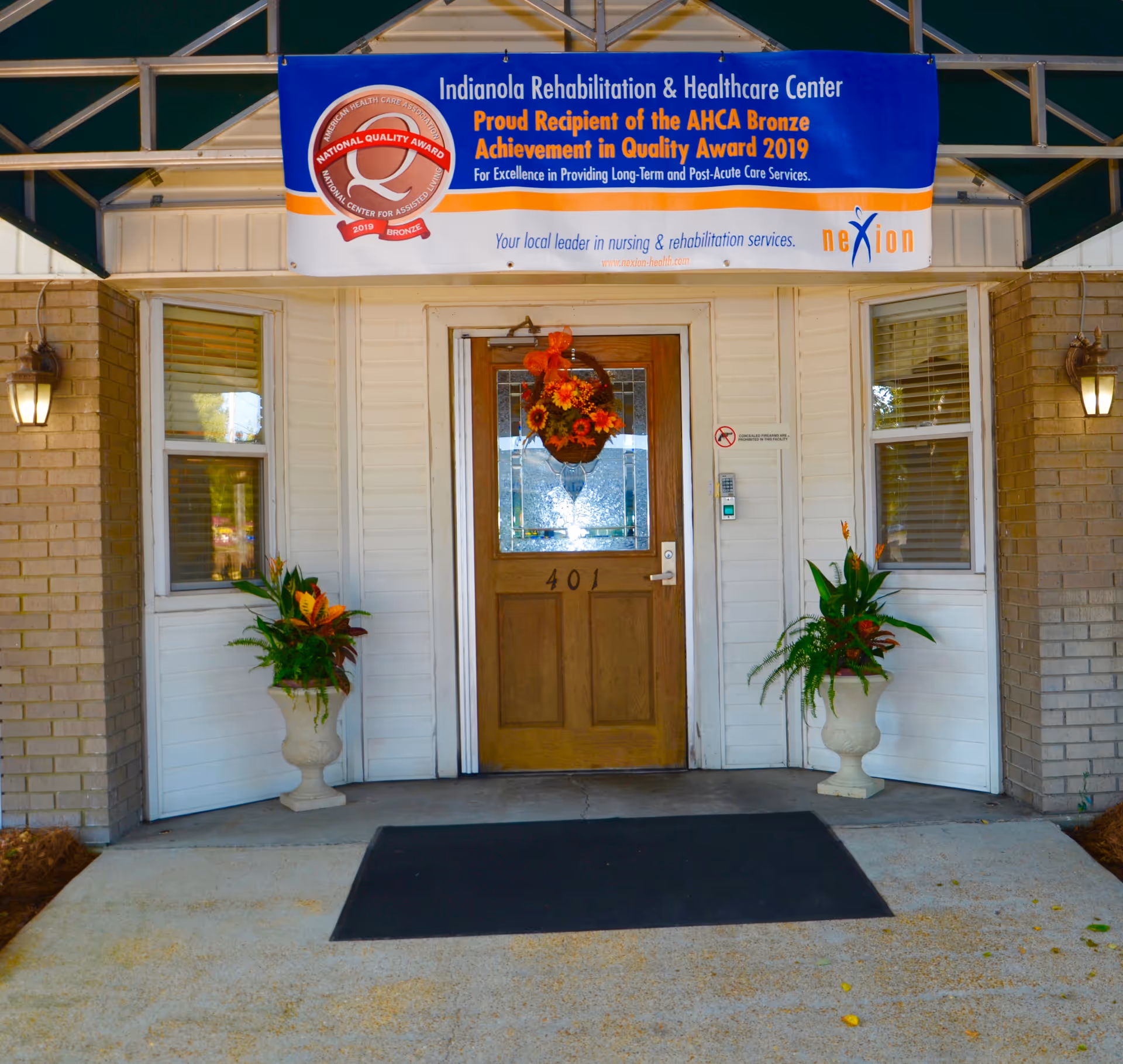 Front entrance of Indianola Rehabilitation & Healthcare Center showing a wooden door numbered 401, potted plants, a wreath, and an award banner above.
