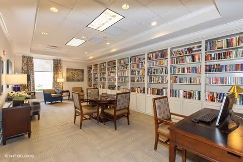 A well-lit room with a large wall of bookshelves filled with books, a central table surrounded by four chairs, a desk with a computer, and comfortable seating near a window with curtains. The room has a carpeted floor and recessed ceiling lights.