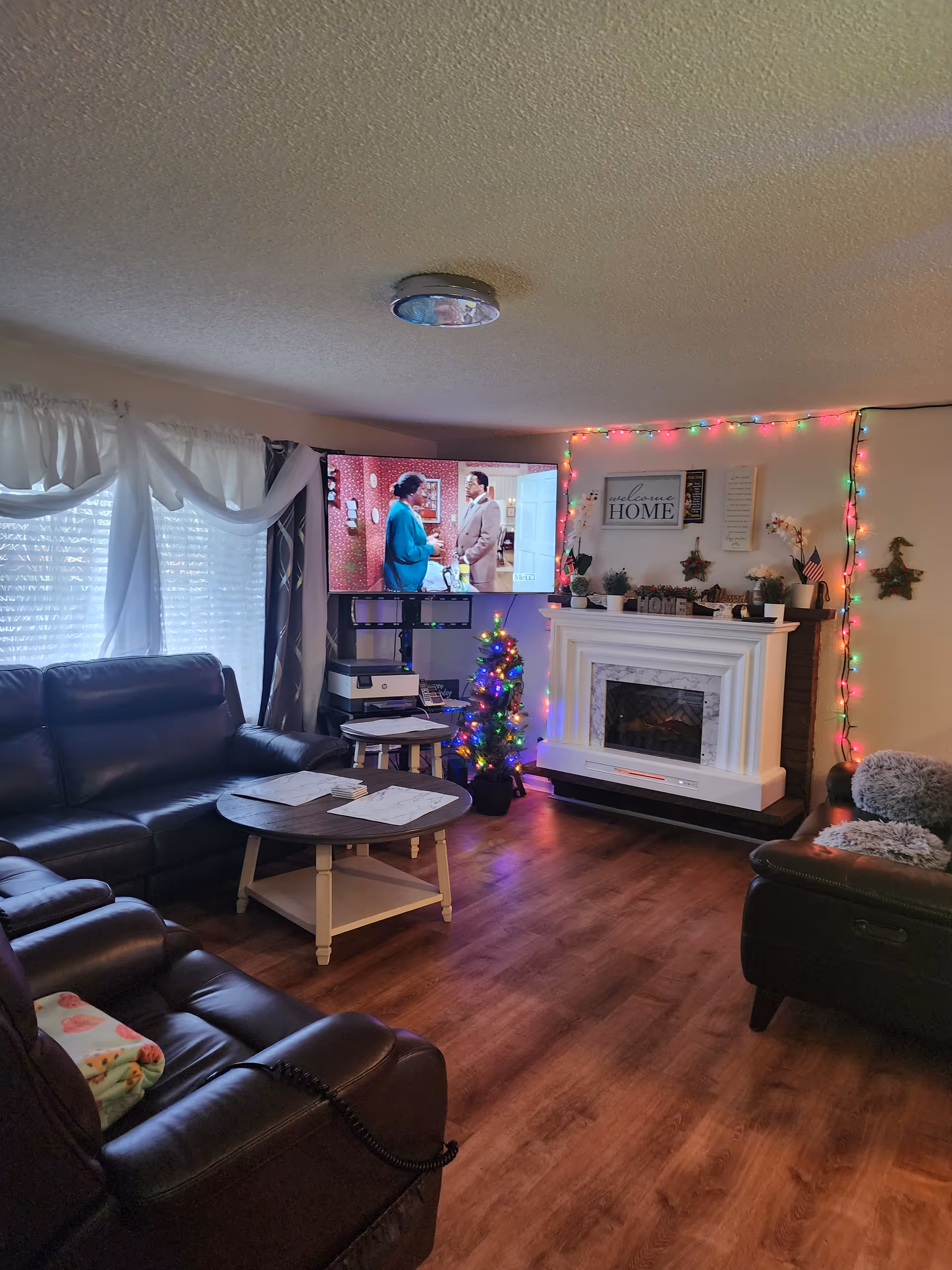 Living room with leather seating, a coffee table, a TV by a small decorated Christmas tree, and a white mantel with holiday lights and decor.