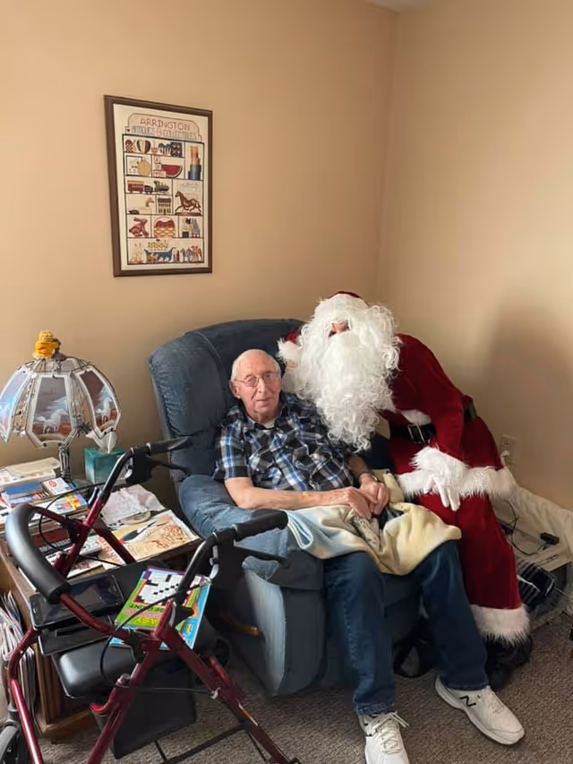 An elderly man sitting in a blue recliner chair with a blanket over his lap, next to a person dressed as Santa Claus who is leaning in close. A red walker is positioned in front of the chair, and a side table with a decorative lamp, books, and other items is nearby. A framed picture hangs on the beige wall behind them.