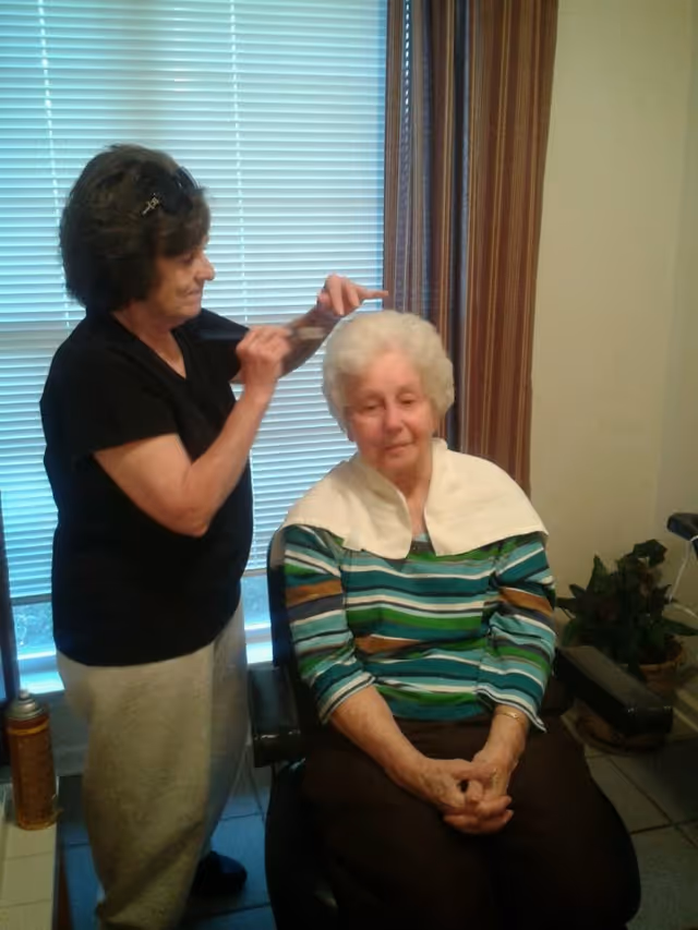 An elderly woman with white hair sits in a chair with a white towel draped over her shoulders while another woman standing beside her styles her hair with a comb. The setting appears to be indoors with a window covered by blinds and striped curtains in the background.