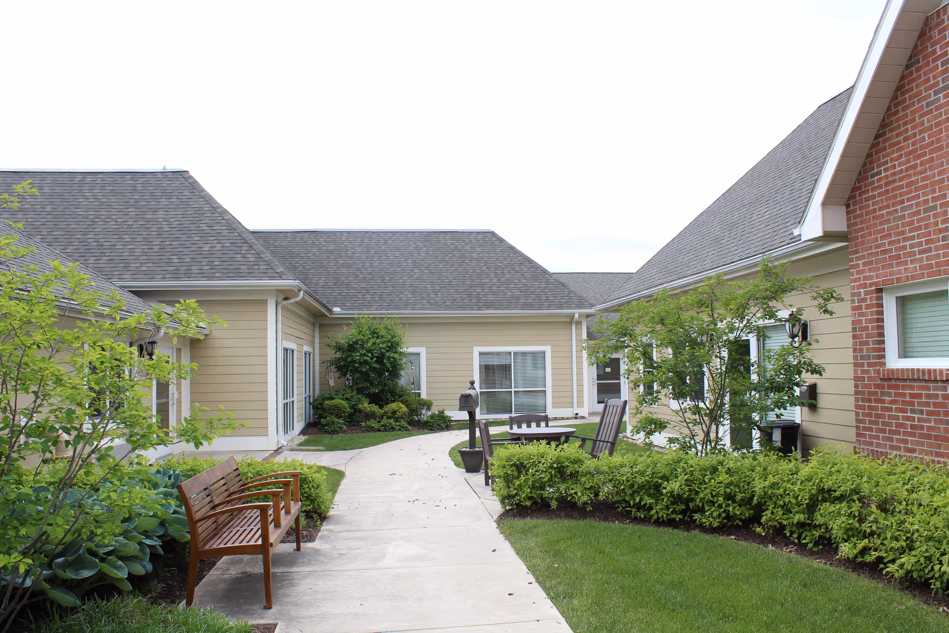 Courtyard with a paved walkway, benches, patio chairs, and landscaped shrubs between single-story senior living buildings.