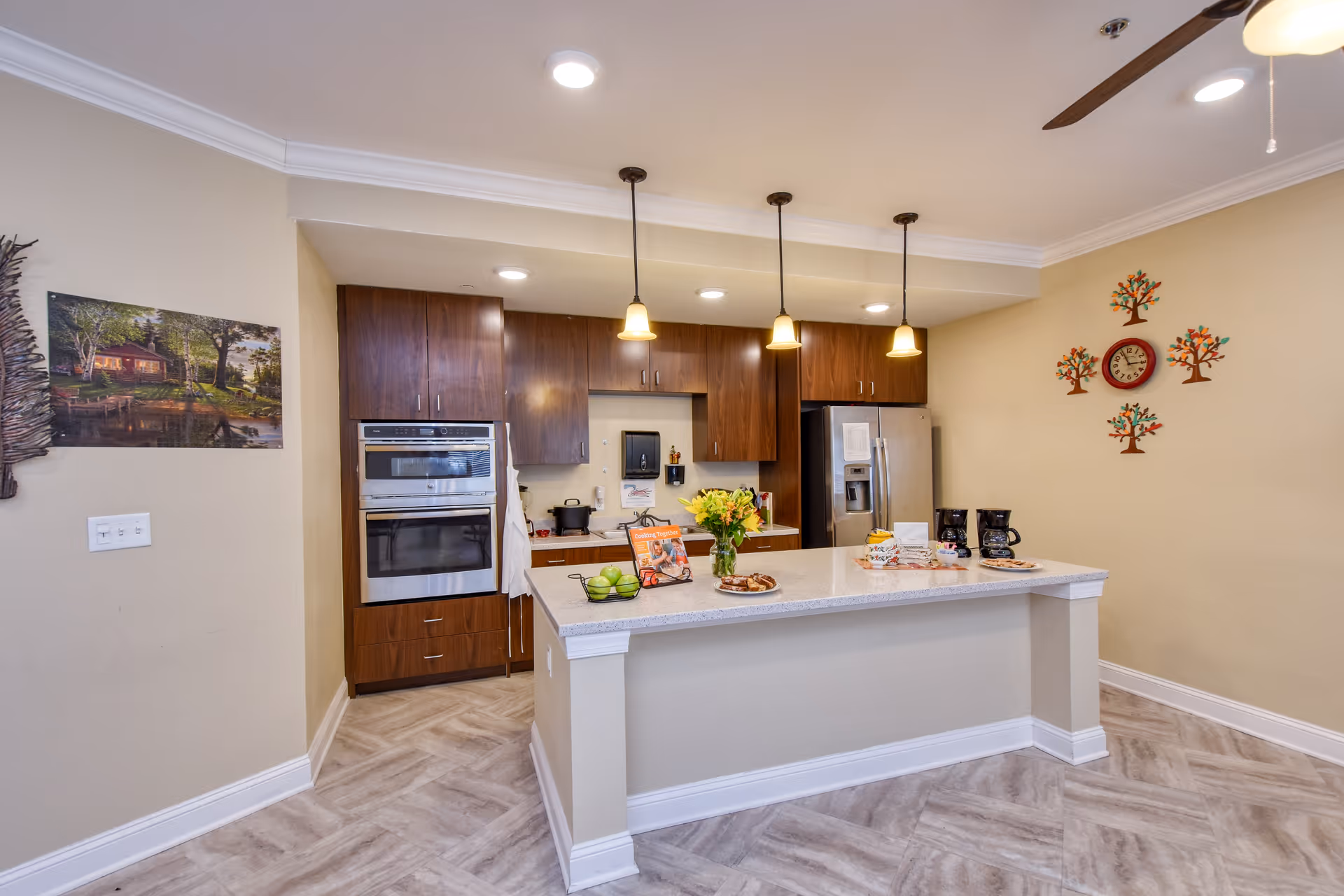 Modern kitchen area with a large island countertop featuring a vase of flowers, a bowl of green apples, and some snacks. The kitchen has wooden cabinets, a double oven, a stainless steel refrigerator, and three pendant lights hanging above the island. The walls are decorated with a clock surrounded by tree-shaped wall art and a landscape painting. The floor has a light-colored herringbone pattern.