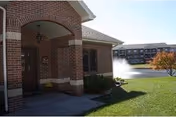 Brick building entrance with an arched doorway, a lantern-style light fixture, and a small flower pot near the door. In the background, there is a pond with a water fountain and a multi-story building with balconies. The scene is set on a sunny day with green grass and a tree with autumn-colored leaves.
