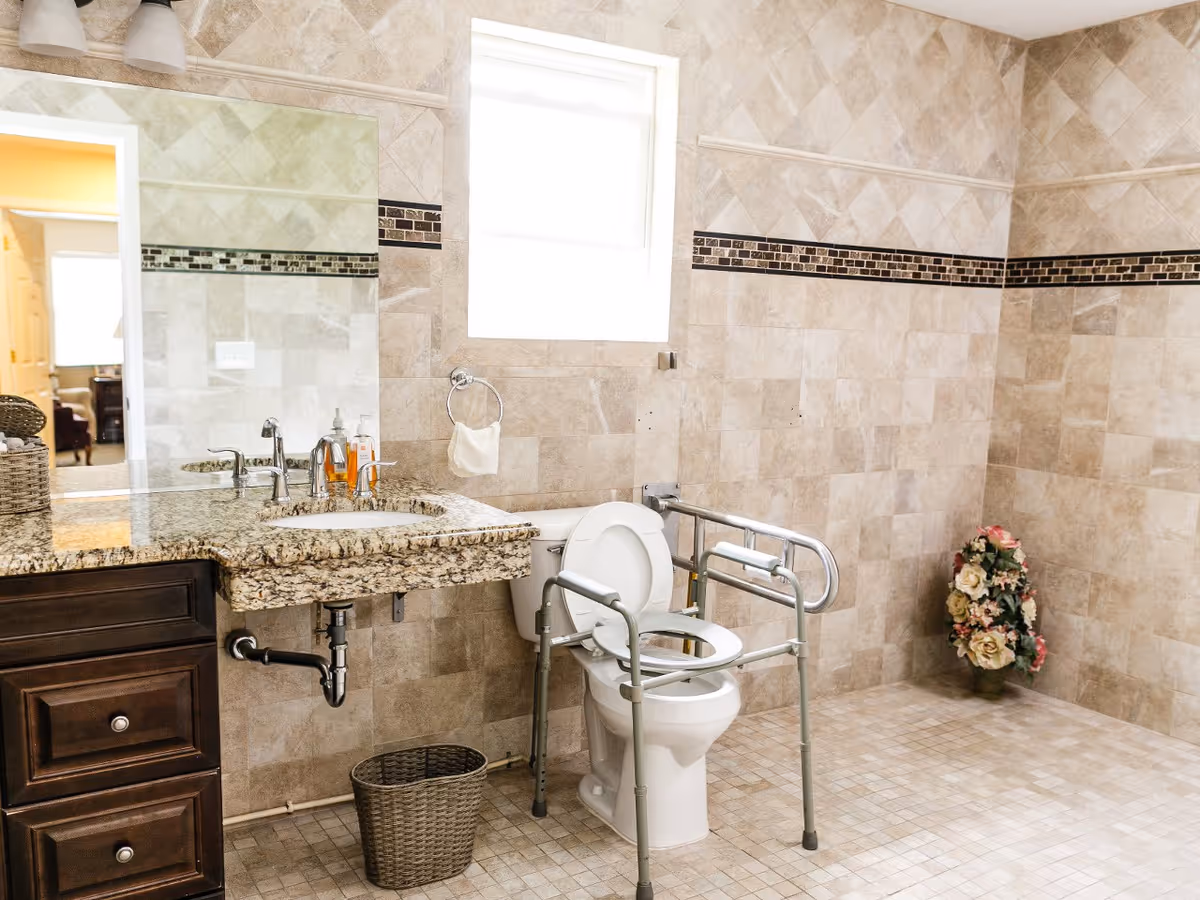 A spacious bathroom with beige tiled walls and floor, featuring a white toilet equipped with safety rails for accessibility, a granite countertop with a sink, soap dispensers, a towel ring with a white towel, a wicker wastebasket, and a floral arrangement in the corner. A large mirror is mounted above the sink, and a window allows natural light into the room.