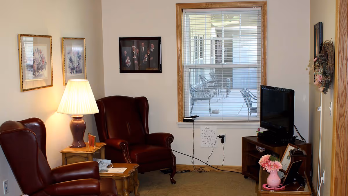 A cozy living room with two dark red leather armchairs, a wooden side table with a lamp, framed pictures on the walls, a window with blinds showing an outdoor patio with chairs, and a wooden TV stand holding a flat-screen television and decorative items including a pink vase with flowers.