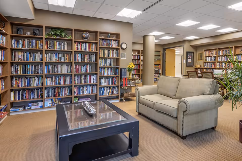 A cozy library or reading room in a senior living facility featuring tall wooden bookshelves filled with books, a beige upholstered sofa, a black coffee table with decorative items, and a carpeted floor. There are also chairs and tables in the background, along with a potted plant and a clock on the wall.