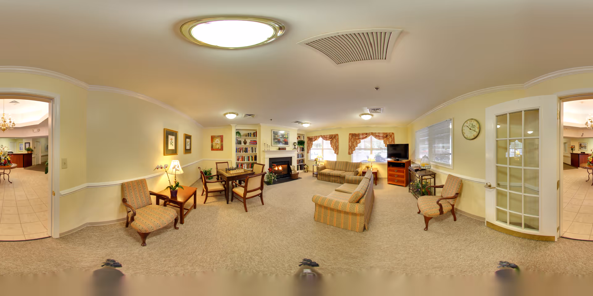 A cozy living room area in a senior living facility with beige walls and carpet. The room features two patterned armchairs, a small wooden table with a lamp and plant, a wooden table with four chairs, a fireplace with a mantel decorated with plants and pictures, a bookshelf filled with books, two large windows with valances, a TV on a wooden stand, and a clock on the wall. French doors and an open doorway lead to other parts of the facility.