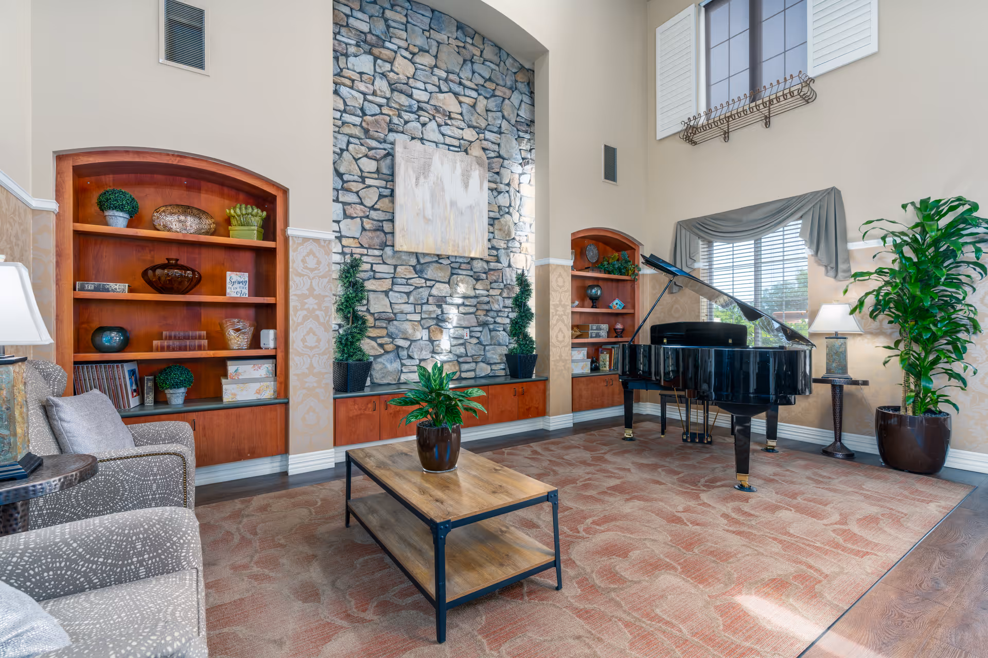 A spacious living room with a grand piano near a window with blinds and a valance. The room features a stone accent wall with a painting, built-in wooden shelves with decorative items, a patterned carpet, a wooden coffee table with a potted plant, and comfortable upholstered chairs. There are also two potted plants and table lamps providing warm lighting.