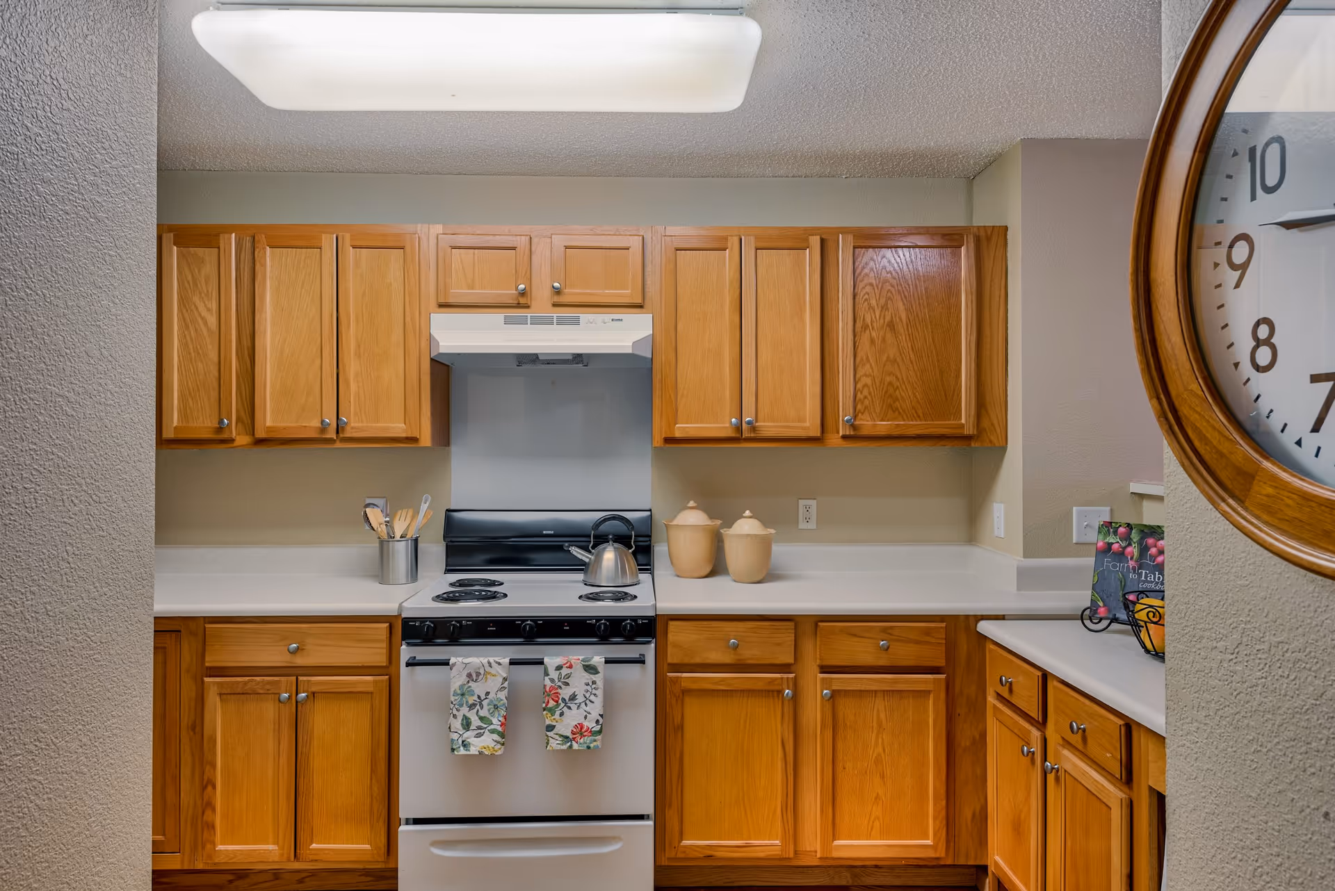 A kitchen with wooden cabinets, a white electric stove with two floral kitchen towels hanging on the handle, a stainless steel kettle on the stove, two beige ceramic containers on the countertop, a utensil holder with wooden utensils, and a large round wall clock partially visible on the right side.