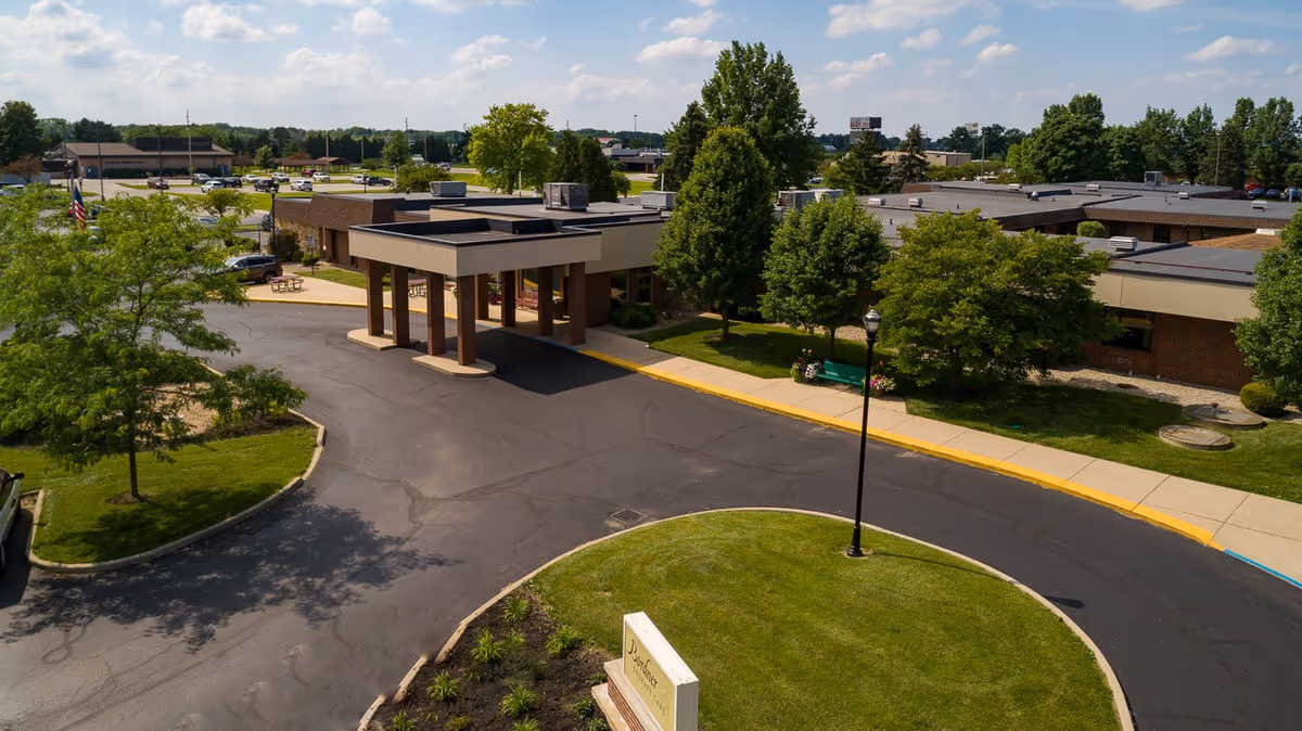 Aerial view of the entrance to Wood Haven Healthcare facility showing a circular driveway with a covered drop-off area, surrounded by green lawns, trees, and parking spaces under a partly cloudy sky.