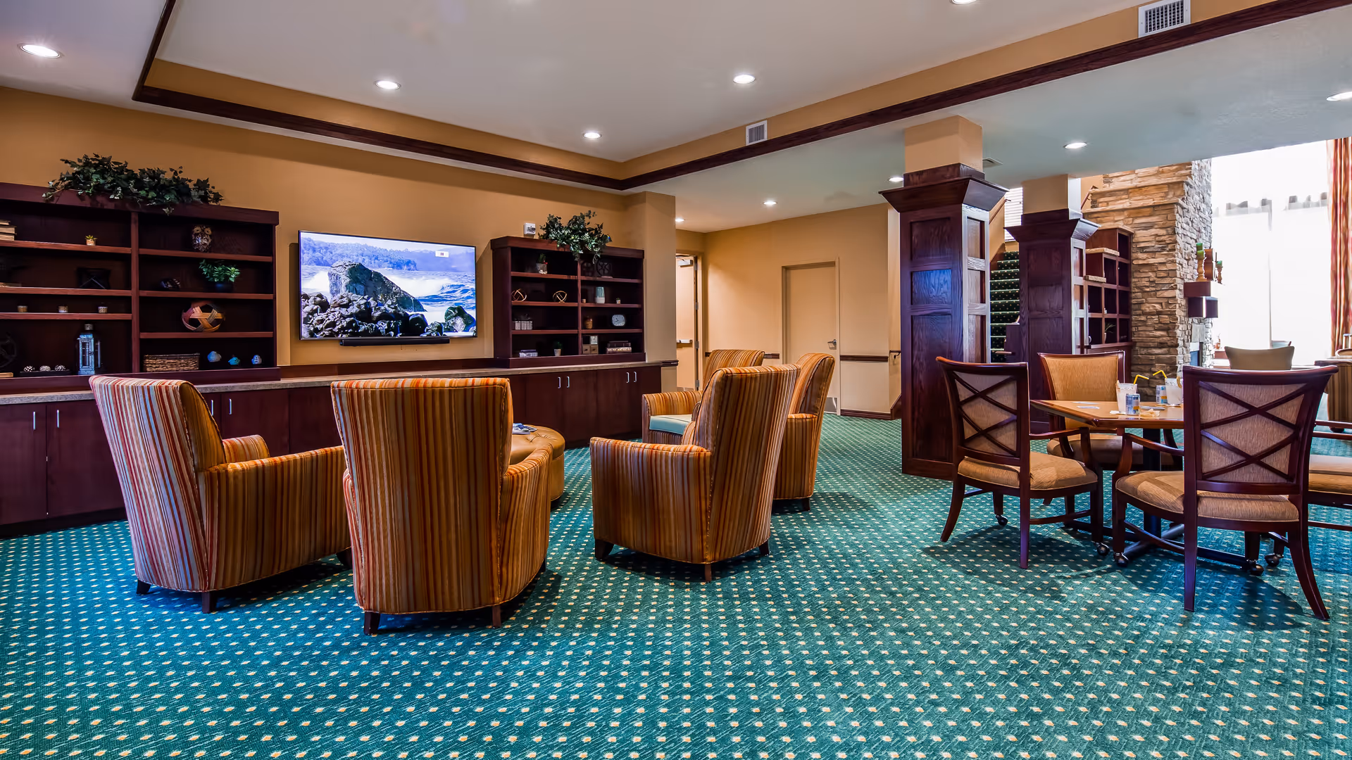 A cozy common area with striped upholstered armchairs arranged around a television mounted on a wall with built-in wooden shelves and cabinets. To the right, there is a round table with four wooden chairs. The room has green patterned carpet, beige walls, and decorative plants on the shelves.