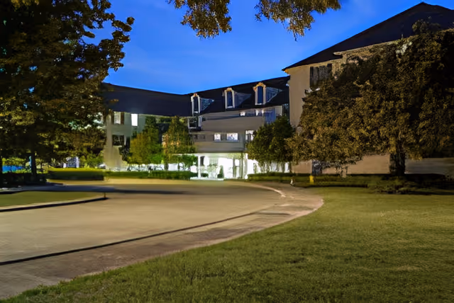 Dusk view of the facility's front entrance with a curved driveway, illuminated entry, lawn and trees.