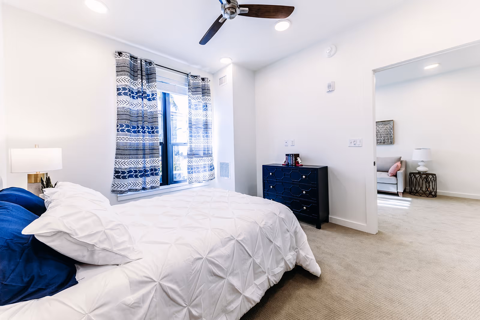 Bright bedroom with a white bedspread and blue pillows, a bedside lamp, patterned blue and white curtains on the window, a dark blue dresser, and an open doorway leading to a living area with a sofa and side table.