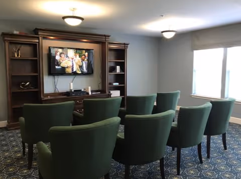 Small common living room with six green upholstered chairs arranged in rows facing a wall-mounted TV in a wooden entertainment unit, patterned carpet, and a window.