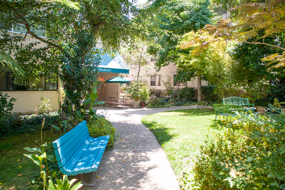 A sunny outdoor garden area with green grass, trees, and bushes. There are two benches, one painted blue and the other green, along a paved pathway. In the background, there is a building with windows and a blue awning labeled 'Stadium' shading a picnic table with an umbrella.