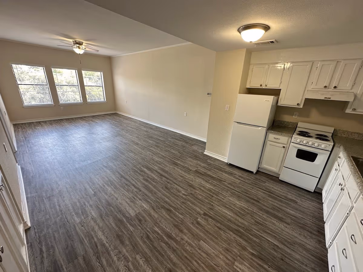 Open-plan living area with wood-look flooring, three large windows and ceiling fan, and a small white kitchen with refrigerator and stove along one wall.