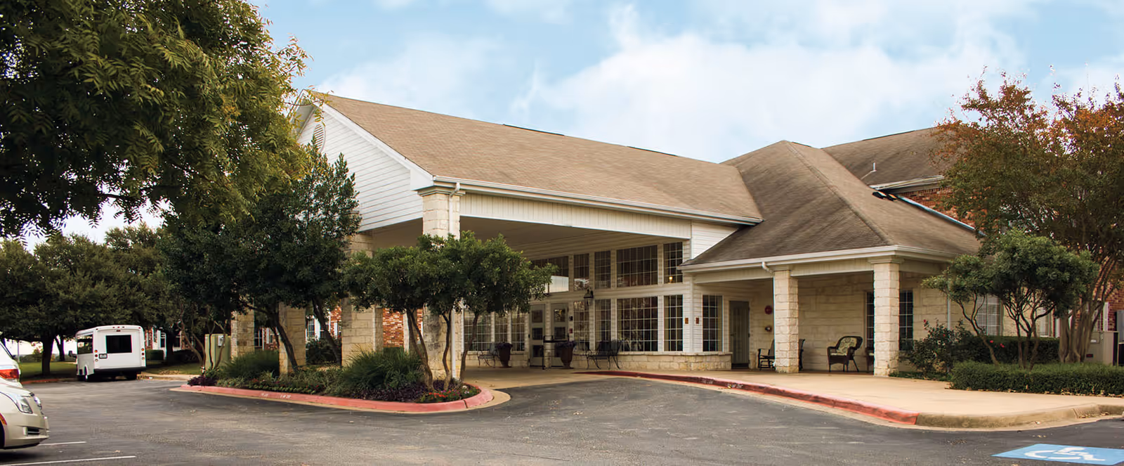 Exterior view of the entrance to Brookdale Round Rock senior living facility, featuring a covered drop-off area with stone pillars, large windows, and surrounding trees and shrubs.