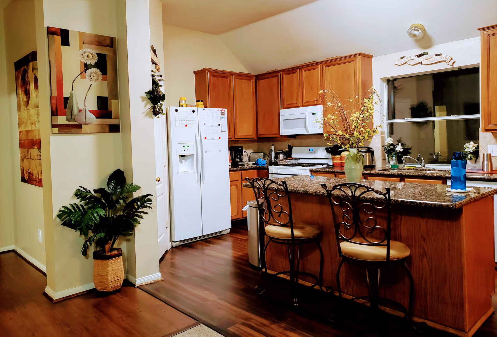 Interior view of a kitchen with wooden cabinets, a white refrigerator, a white microwave, and a stove. There is a kitchen island with a granite countertop and two decorative chairs. A green vase with flowers is placed on the island. The floor is wooden, and there are framed artworks and a potted plant near the entrance.
