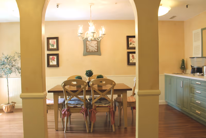 Dining room with a wooden table and chairs under a chandelier, decorative wall art, and adjacent mint-green kitchen cabinets.