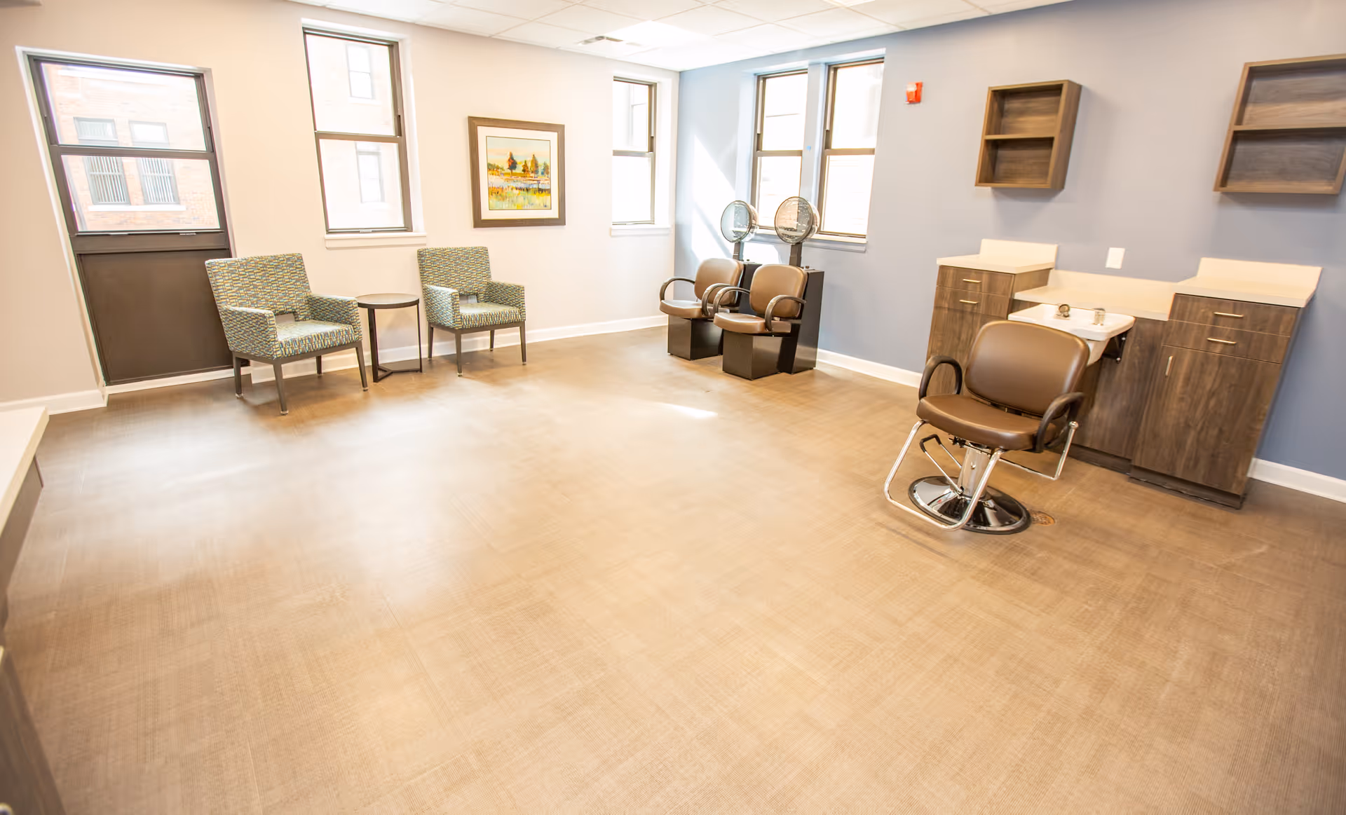 A bright and spacious hair salon area in Norman Towers Senior Apartments featuring a styling chair in front of a sink with cabinets, two hair drying chairs with hooded dryers, two patterned armchairs with a small round table between them, and three windows letting in natural light.