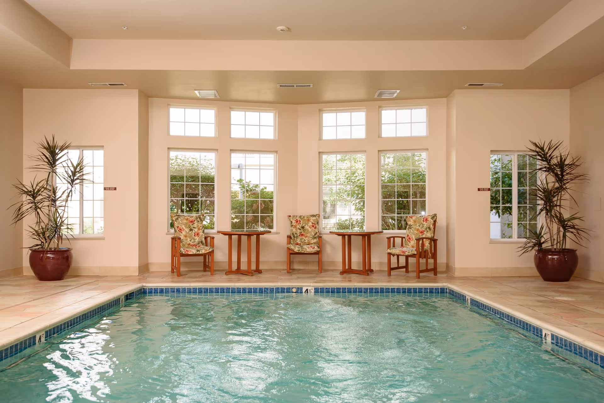 Indoor swimming pool with clear water, surrounded by beige tiled flooring. Along the far wall, there are three wooden chairs with floral cushions and two small wooden tables between them. Large windows behind the chairs let in natural light and show greenery outside. Two large potted plants are placed on either side of the room.