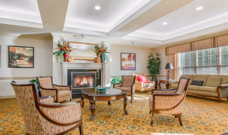 A cozy senior living community lounge with a lit fireplace, four patterned armchairs arranged around a wooden coffee table, a beige sofa, an armchair with a red pillow, large windows with blinds, decorative plants, and framed artwork on the walls.