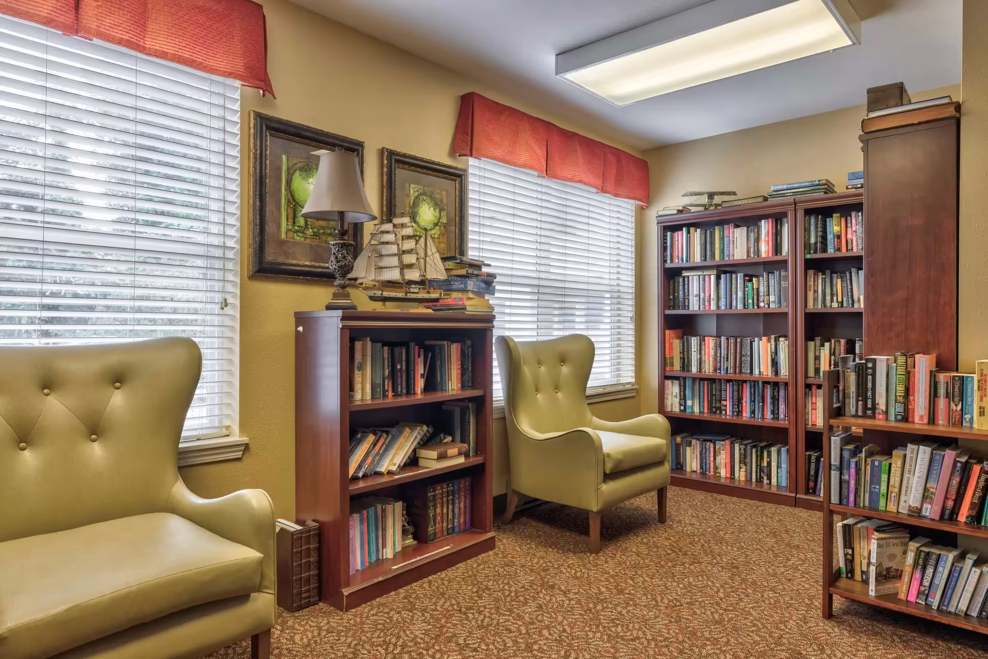 A cozy reading room with two green upholstered armchairs, several wooden bookshelves filled with books, a table lamp, and decorative framed artwork on the wall. The room has two windows with white blinds and red valances, and a patterned carpeted floor.