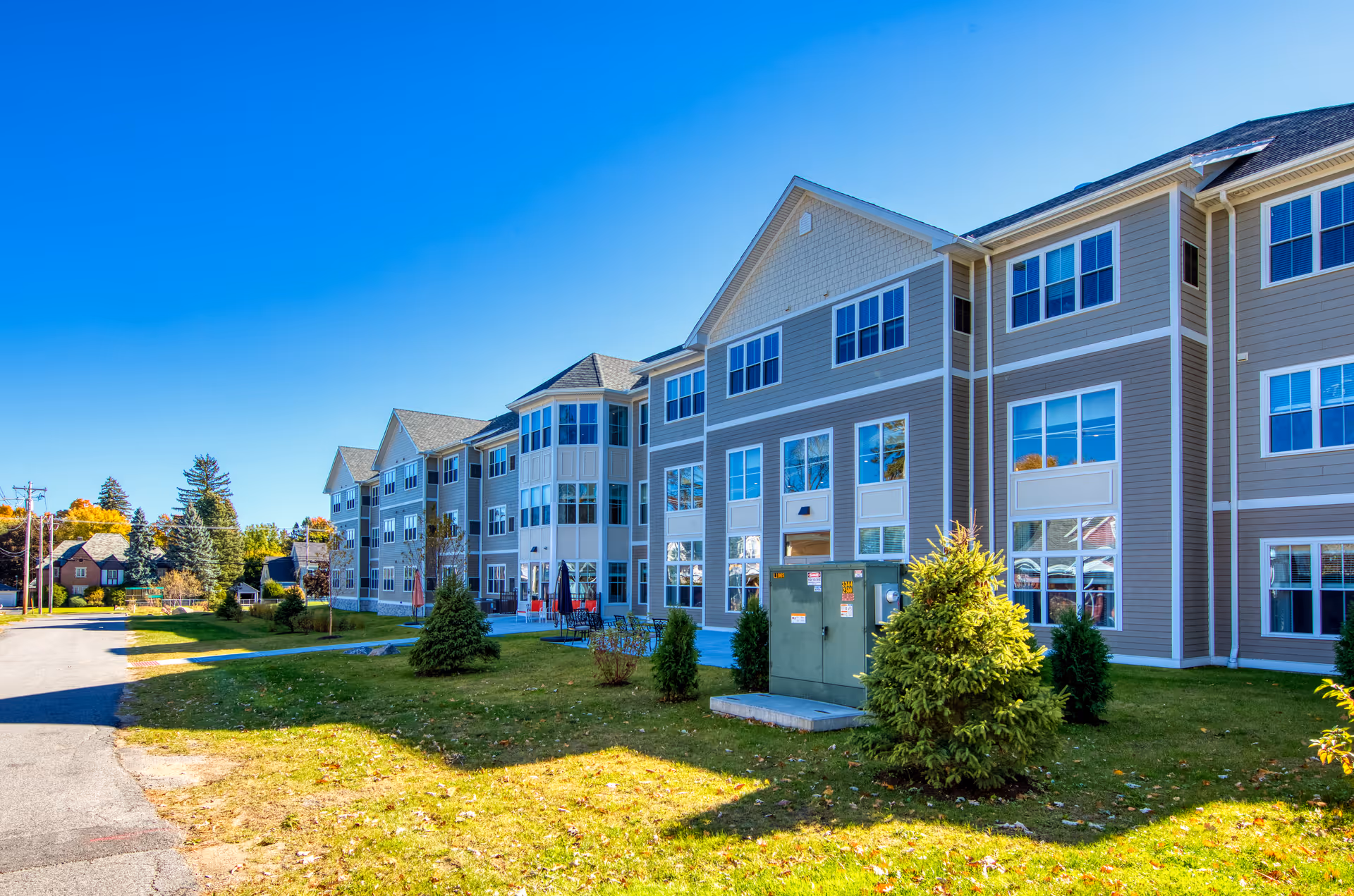 Exterior view of a large, multi-story senior living facility building with beige siding and many windows under a clear blue sky. There is a grassy area with small trees and shrubs in front of the building, and a paved road runs alongside it.