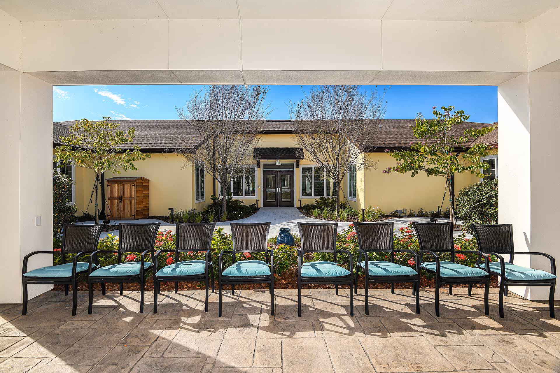 Row of patio chairs with teal cushions under a covered walkway facing a landscaped courtyard and the entrance to a single-story building.