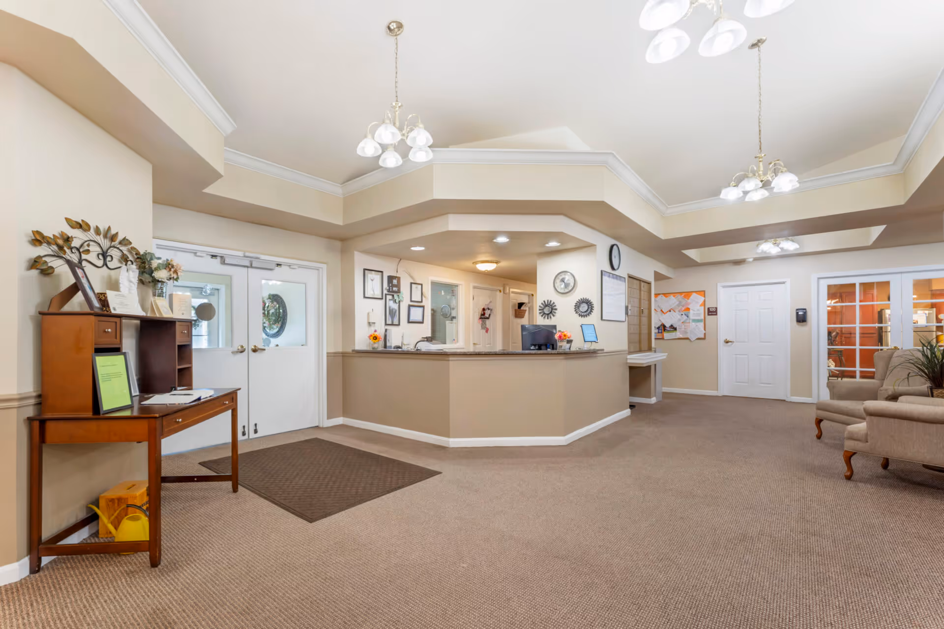 Reception area of The Addison of McCullough Place featuring a beige carpeted floor, a front desk with computer monitors, wall clocks, and framed pictures. There are double doors with round windows on the left, a wooden table with decorative items and documents, and a seating area with beige armchairs on the right. The ceiling has recessed lighting and hanging light fixtures.