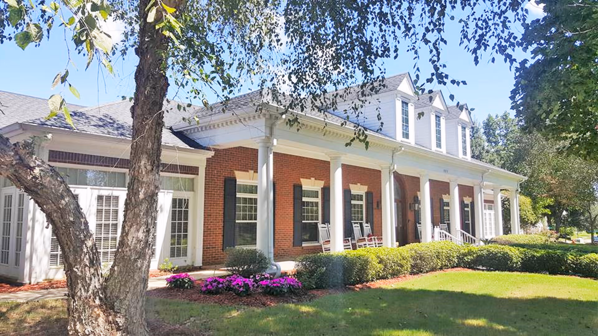 Brick senior living building with a columned front porch, rocking chairs, landscaped lawn and trees.