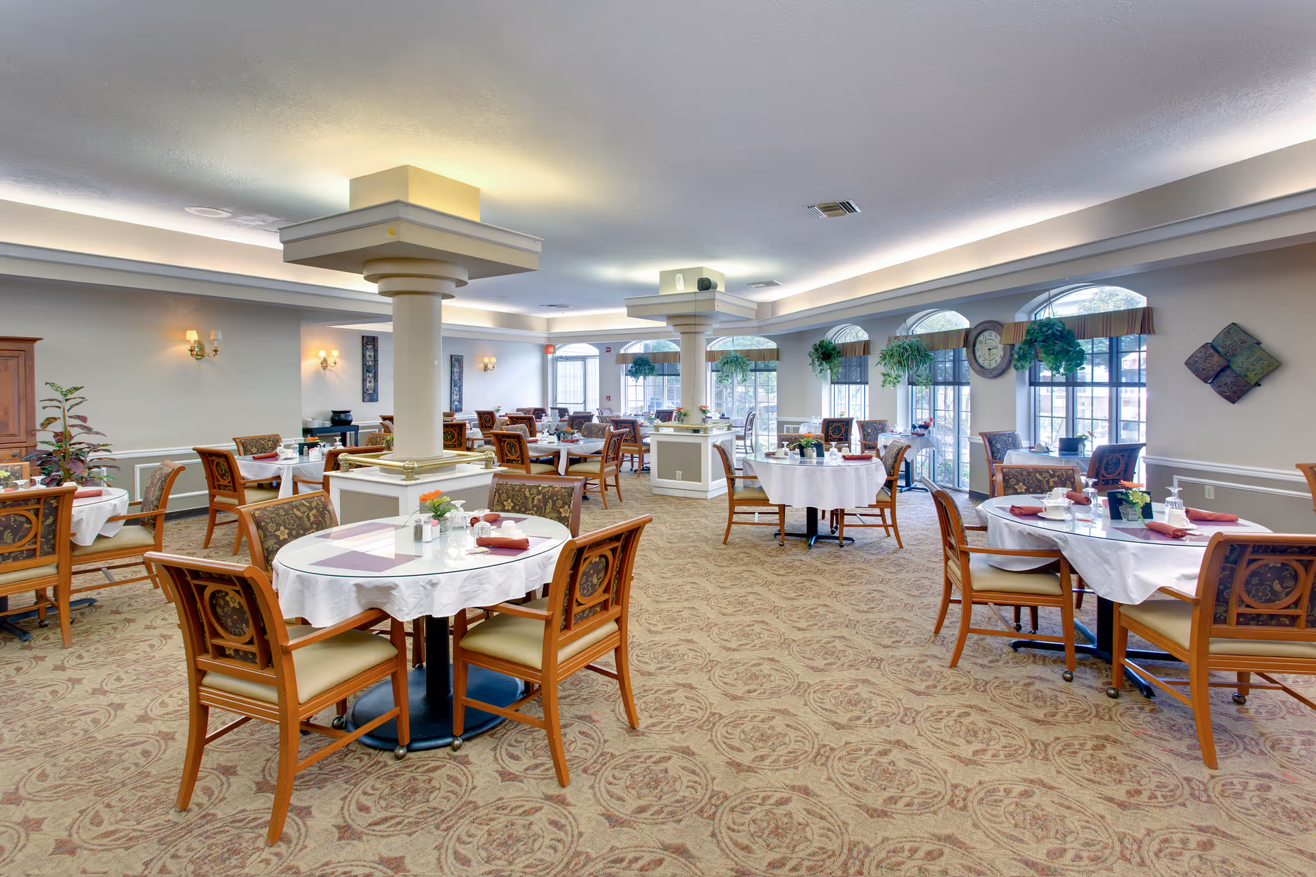 A spacious dining room with multiple round tables covered with white tablecloths and set with placemats, napkins, and small flower arrangements. The room has patterned carpet, large windows with arched tops letting in natural light, hanging plants, and decorative wall art. Two large square columns are centrally located in the room with soft ceiling lighting above them.
