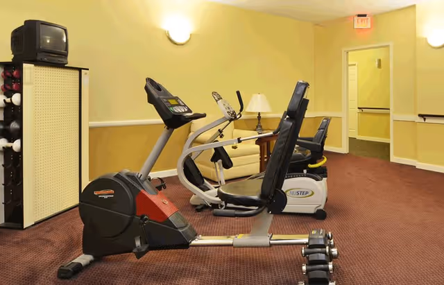 Indoor exercise room with two recumbent stationary bikes, a small TV on a stand, a rack of dumbbells, a beige couch, a side table with a lamp, and yellow walls with wall-mounted lights.