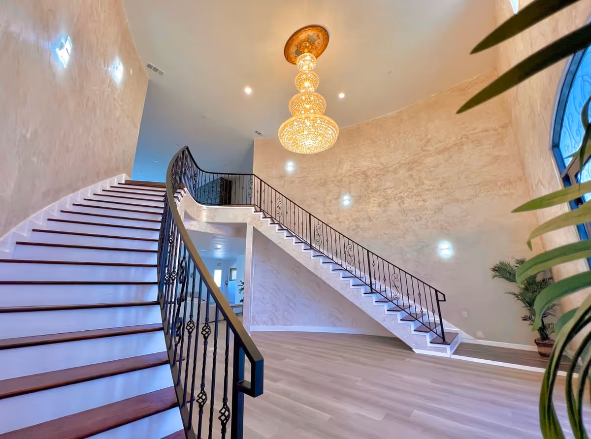 Interior view of a spacious area with a curved staircase featuring dark wooden steps and black wrought iron railing. The walls have a textured beige finish and are adorned with small wall lights. A large, ornate chandelier hangs from the ceiling. There is a large arched window on the right side with some green plants nearby. The floor is light wood.