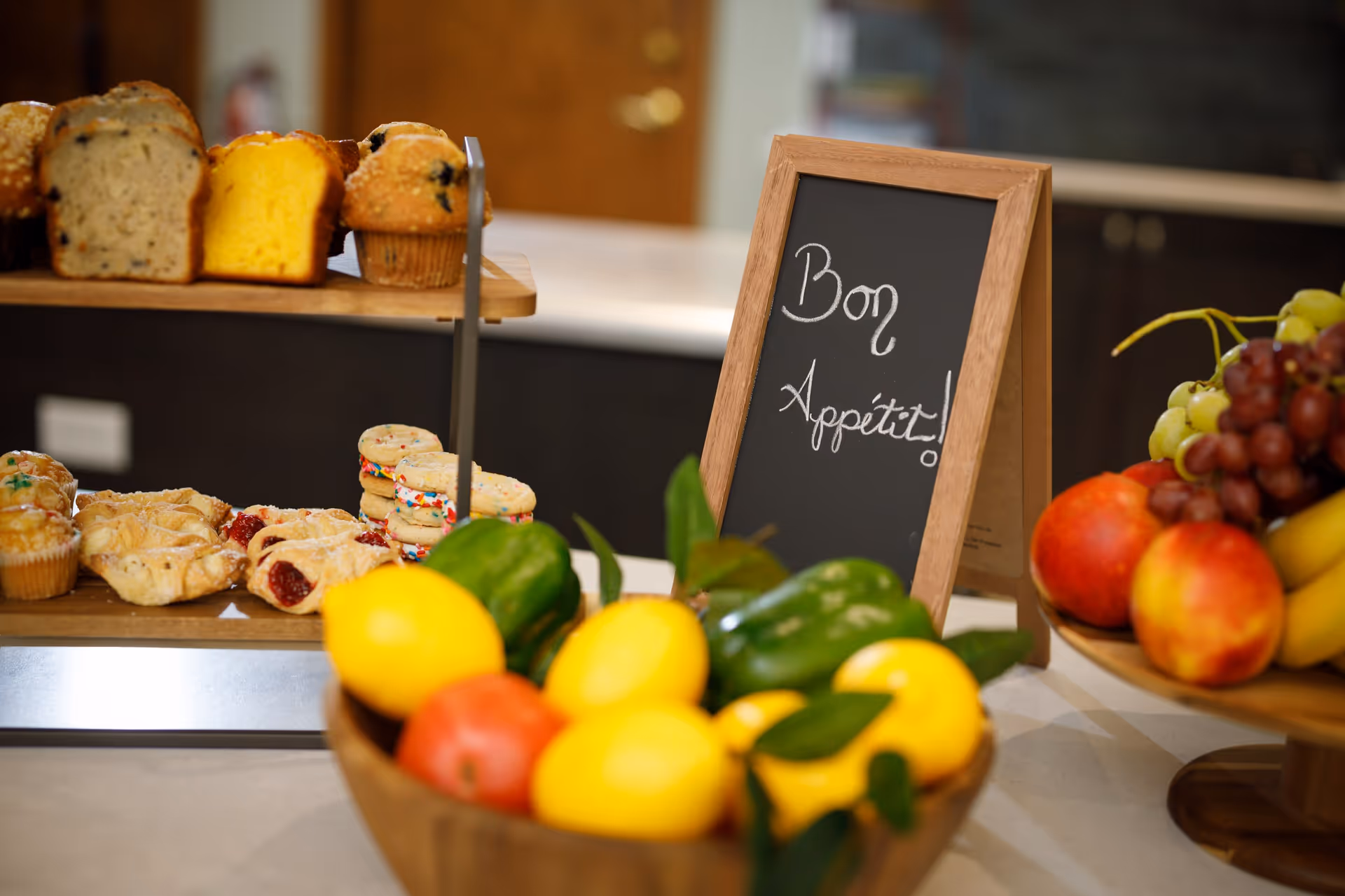 A kitchen counter displaying a variety of baked goods including muffins, bread, cookies, and pastries on a wooden tiered stand. In front, there is a wooden bowl filled with fresh fruits and vegetables such as lemons, green bell peppers, and apples. To the right, a small chalkboard sign reads 'Bon Appétit!'.