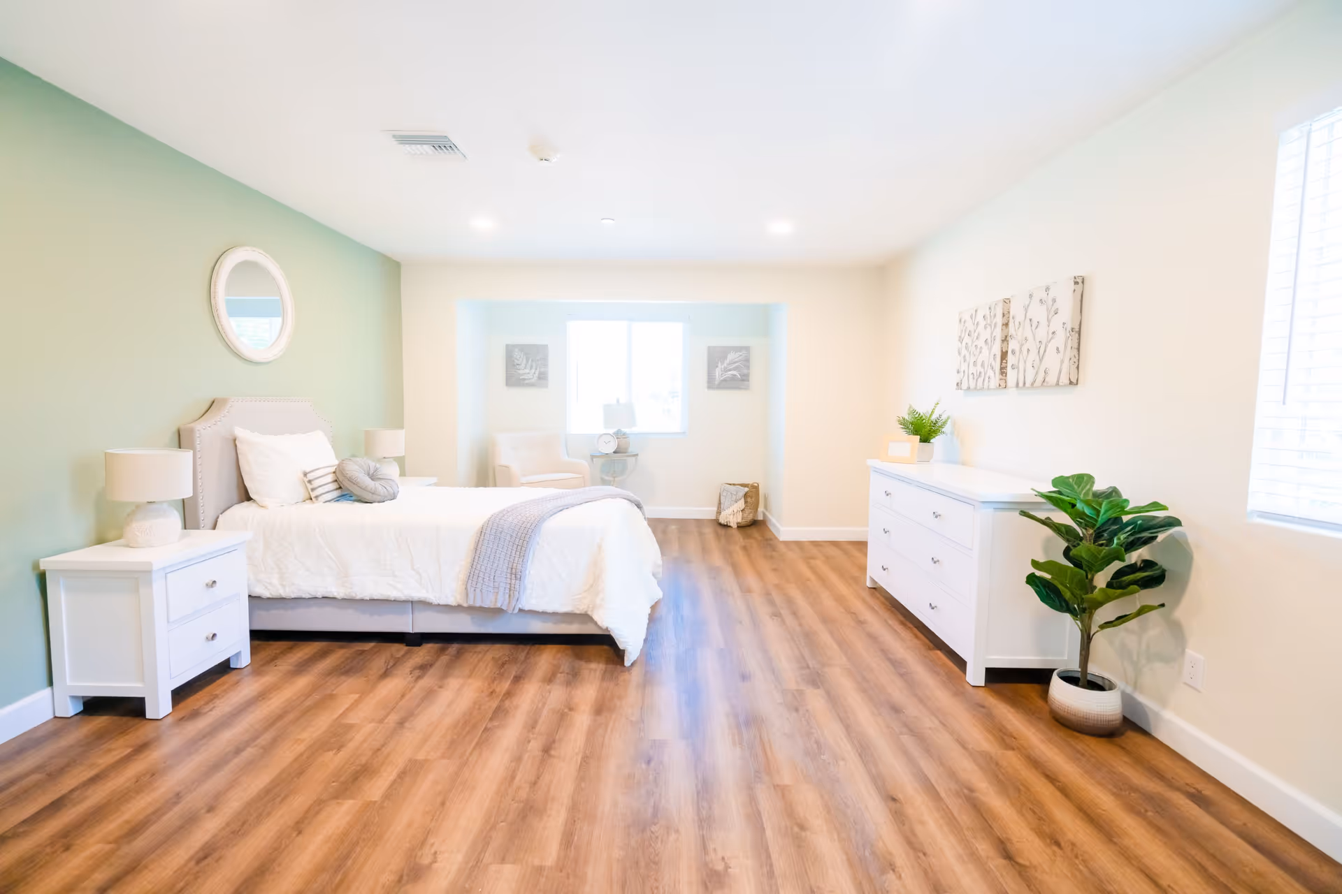 A bright and spacious bedroom with light wood flooring, a bed with white bedding and pillows, two white nightstands with lamps, a white dresser with decorative items, a green accent wall with a round mirror, a window with white blinds, a small sitting area with a chair and side table, and a potted plant in the corner.