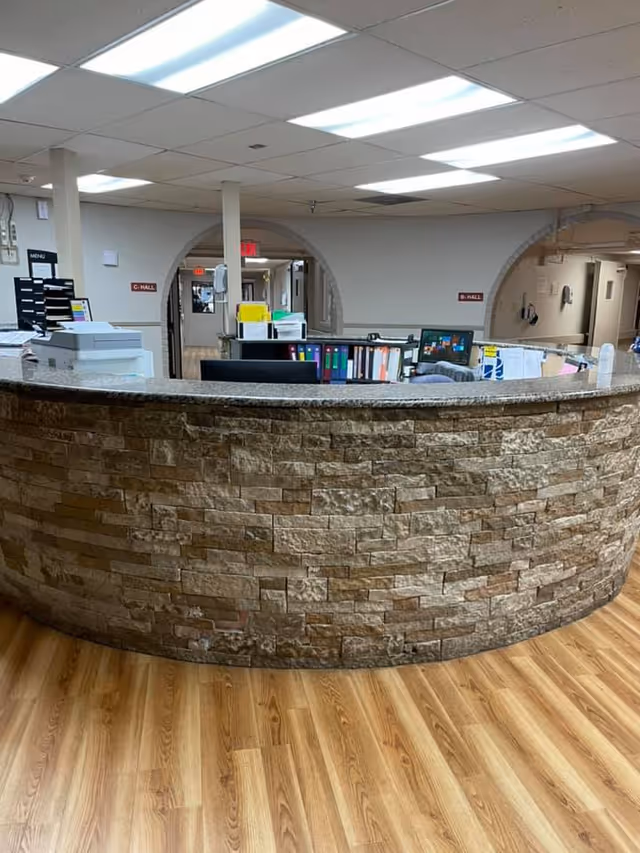 Curved reception desk with a stone facade in a nursing and rehabilitation center. The desk has office supplies, folders, and computer monitors on it. Behind the desk, there are hallways labeled C-Hall and B-Hall with arched doorways. The floor is wood-patterned, and the ceiling has fluorescent lighting.